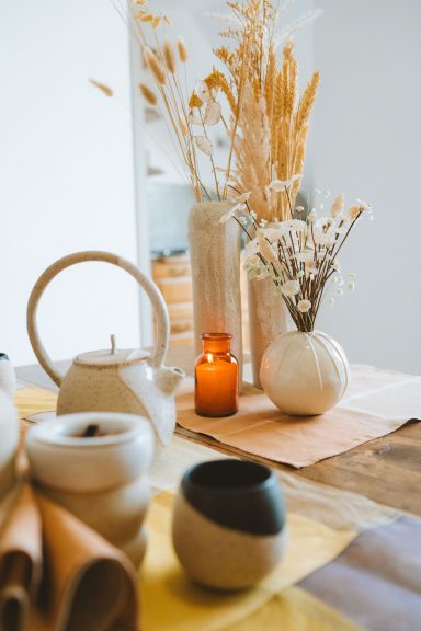 Vases avec des fleurs sèches, bougie orange et vaisselle sur une table en bois.