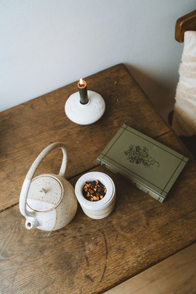 Théière blanche, bougie allumée, pot de fleurs et boîte vintage sur une table en bois.